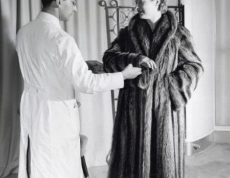 Een vrouw [in bontwinkel] bezig met het passen van een jas. De bontcoupeur inspecteert de bontjas / bontmantel van de cliënte. De jas bestaat uit 34 wasbeerhuidjes. Nederland, Amsterdam, 21 augustus 1953. Foto: Wout van de Hoef