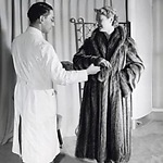 Een vrouw [in bontwinkel] bezig met het passen van een jas. De bontcoupeur inspecteert de bontjas / bontmantel van de cliënte. De jas bestaat uit 34 wasbeerhuidjes. Nederland, Amsterdam, 21 augustus 1953. Foto: Wout van de Hoef